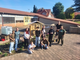 Auf einem gepflasterten Hof mit Grünstreifen stehen zehn junge Menschen vor einem Insektenhotel.