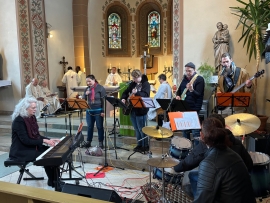 Im Altarraum einer Kirche sind im Vordergrund Musiker einer Band mit Piano, Schlagzeug, Gesang und Gitarren. Im Hintergrund am alter stehen drei Priester in weißen Gewändern.