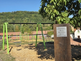 Ein Spielgerüst auf einer Wiese, im Hintergrund ein Waldhügel und im Vordergrund eine Holzstele mit Plakette
