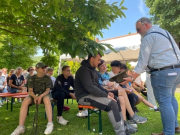 Eine Gruppe Jugendlicher in sommerlicher Kleidung sitzt auf Bierbänken unter einem Baum. Ein Mann mit Hut geht durch die Reihen.