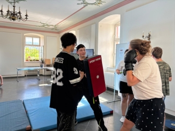 Eine junge Frau mit Boxhandschuhen steht vor zwei Jugendlichen, die eine Pratze halten.