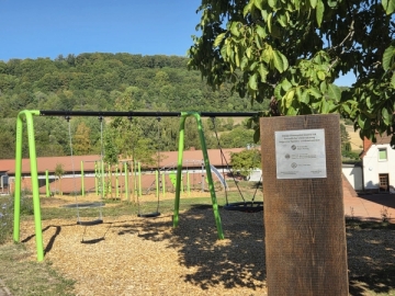 Ein Spielgerüst auf einer Wiese, im Hintergrund ein Waldhügel und im Vordergrund eine Holzstele mit Plakette