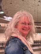 Eine Frau sieht in die Kamera. Sie tr&auml;gt eine Brille, hat graues langes Haar und ist mit rosa Schal und einer Jeansjacke bekleidet.