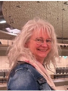 Eine Frau sieht in die Kamera. Sie tr&auml;gt eine Brille, hat graues langes Haar und ist mit rosa Schal und einer Jeansjacke bekleidet.