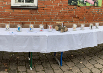 Draußen vor der Einrichtung stehen Gläser mit bunt gestalteten Kerzen stehen aufgereiht auf einem weiß gedeckten Tisch.